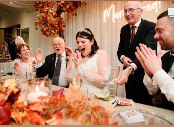 top table at wedding. bride and grrom wiht close fmaily ina  loine. bride is open mouithed with surprise at the revelation of a trick